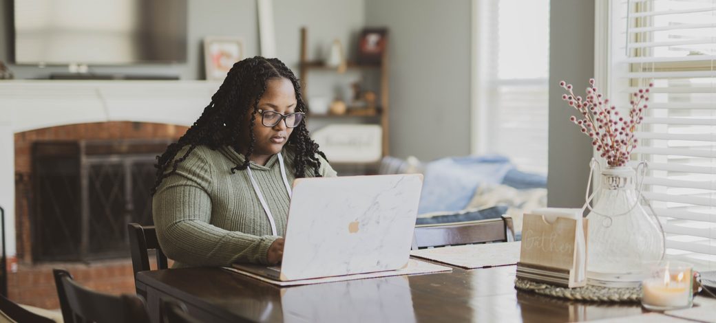 A Black woman with long braids and glasses works on a laptop at her dining room table at home.